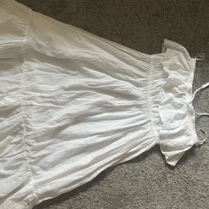 Vit somrig långklänning med volang - Superfin vit långklänning med axelbandslös design och volang upptill. Klänningen har smock i midjan för en bekväm passform och är gjord i ett lätt bomullstyg, perfekt för varma dagar. Enkel och stilren look som känns fräsch och luftig.