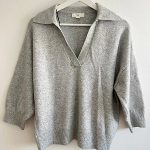 Grå stickad polotröja från H&M - Mysig grå stickad polotröja från H&M med v-ringning och bred krage. Tröjan har ribbade muddar och avslut, samt en loose passform som ger en avslappnad vibe. Perfekt för lager-på-lager under kyliga dagar.