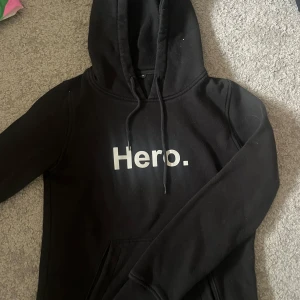 Svart hoodie med Hero-tryck - Svart hoodie med vit text 'Hero.' på bröstet. Klassisk modell med huva och snörning samt magficka. Perfekt för dig som gillar enkel och stilren streetwear.
