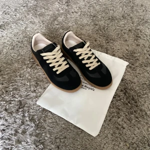 Svarta sneakers Maison Margiela - Stilrena svarta sneakers från Maison Margiela med beige snörning och gummisula i naturfärg. Ovandelen kombinerar skinn och mocka för en lyxig känsla. Klassisk rund tå och platt sula gör dem perfekta för en trendig vardagslook.