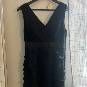 Svart klänning med volanger - Snygg svart klänning med v-ringning och breda axelband. Klänningen har flera lager volanger nedtill och omlottliknande överdel. Perfekt för dig som gillar en klassisk men ändå trendig look. Aldrig använd så i topp skick! Passar mig som har XS i kläder men också min mamma som har M/L så passar dem flesta😊