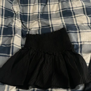 Svart volangkjol från Quzu - Supersöt svart kortkjol från Quzu med smockad midja och volangkant. Kjolen har en flowy känsla och är perfekt för dig som gillar en feminin och trendig stil. Materialet är lätt och ger en snygg siluett💕💕