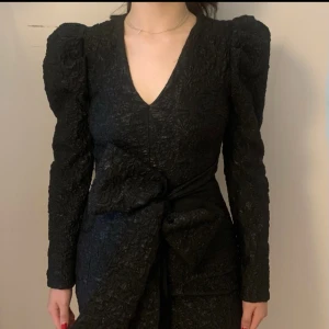 Svart kort klänning med puffärm och rosett - Supersnygg svart kort klänning med markerade axlar, puffärmar och v-ringning. Klänningen har ett coolt strukturerat tyg med blommönster och en stor rosett i midjan. Perfekt för dig som vill sticka ut med en unik look. Använd en gång stl 36 pris 250kr