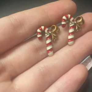 Juliga örhängen med polkagrisar - Söta örhängen formade som polkagrisstavar med rödvita ränder och en liten guldfärgad rosett. Perfekta för att maxa julkänslan och sticka ut med en lekfull detalj. Stift i metall och färgglad emaljfinish.