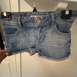 Levi's ljusblå jeansshorts - Snygga ljusblå jeansshorts från Levi's med klassisk femficksdesign, uppvikta benslut och slitna detaljer. Shortsen har normal passform och är perfekta för varma sommardagar. Ikonisk Levi's-lapp bak och röd etikett på bakfickan.