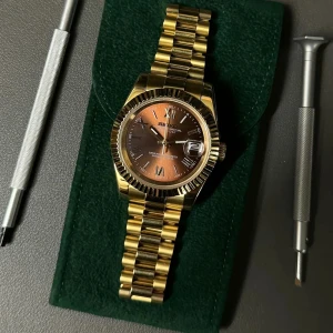 Seiko Datejust automatisk guld klocka - Snygg Seiko Oyster Perpetual Datejust med automatisk urverk, guldigt armband och boett. Urtavlan har en cool brun ton med romerska siffror och datumfönster vid kl 3. Länken är robust och glänsande, perfekt för dig som gillar statement-accessoarer.