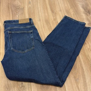 Blå jeans från Dressmann relaxed fit - Snygga blå jeans från Dressmann i relaxed fit med klassisk femficksdesign och raka ben. Jeansen har en mörkblå tvätt, kontrastsömmar och är tillverkade i slitstarkt bomullsjeansmaterial. Perfekta för en chill och avslappnad stil.
