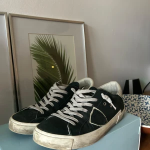 Philippe Model mörkblåa sneakers i mocka - Snygga mörkblåa Philippe Model sneakers i mocka med vita detaljer och grå snörning. Skorna har vit sula med vintage-look och vit hälkappa med logga. Klassisk låg modell med rund tå och coola metalldetaljer vid snörningen.