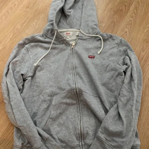 Grå hoodie med dragkedja från Levi's - Säljer en grå hoodie från Levi's i storlek S. Tröjan har dragkedja framtill, huva med snörning och klassisk röd Levi's-logga på bröstet. Tillverkad i mjuk bomullsmix, perfekt för chill dagar. Fickor framtill för extra bekvämlighet.