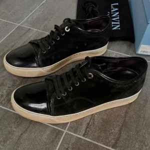 Svarta sneakers från Lanvin - Stilrena svarta sneakers från Lanvin med ovandel i mocka och lackad tå. Skorna har svarta snören, rund tå och vit sula som ger en snygg kontrast. Perfekta för dig som gillar en clean och lyxig look.