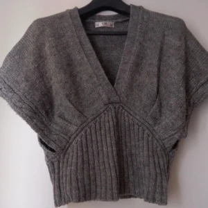 Grå stickad tröja från Golden Days - Kortärmad grå stickad tröja från Golden Days med djup v-ringning och bred ribbad midja. 