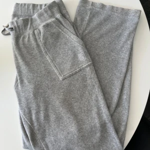 Grå mjukisbyxor från Juicy Couture - Grå mjukisbyxor från Juicy Couture med dragsko i midjan, stora fickor och diskret logga framtill. Byxorna är i mjukt bomullsmaterial och har snygga detaljer med knappar på bakfickan. Perfekta för chill dagar eller när du vill vara extra comfy.