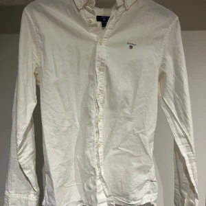 Vit skjorta från GANT med broderad logga - Klassisk vit skjorta från GANT med broderad logga på bröstet. Skjortan har lång ärm, knappar hela vägen och en snygg krage. Tillverkad i bomull för en skön känsla och stilren look. Perfekt för dig som gillar enkel och tidlös stil. Skjortan är i bra skick! Bara att höra av sig vid eventuella frågor!😉