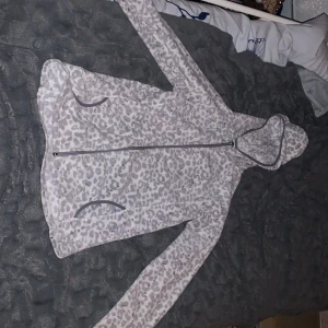 Leopardmönstrad fleecejacka med huva - Mysig fleecejacka med grått leopardmönster och hel dragkedja framtill. Jackan har huva och två fickor, perfekt för chill dagar. Långärmad och i mjukt material som håller dig varm. Och vi kan prata om priset om det behövs! 
