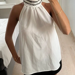 Vit halterneck blus med pärldetaljer - Säljer en vit halterneck blus med hög krage dekorerad med svarta pärlor och broderade detaljer. Toppen är ärmlös och har en luftig, flowy passform som ger en snygg siluett. Perfekt för dig som vill sticka ut med coola detaljer och elegant look.