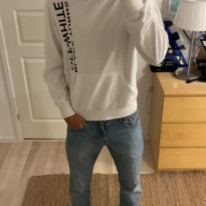 Vit Off-White hoodie med tryck - Säljer en vit hoodie från Off-White med svart texttryck på framsidan och stort grafiskt tryck i svart och silver på ryggen. Klassisk känguruficka, huva och ribbade muddar. Perfekt för dig som gillar streetwear och vill sticka ut.