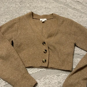 Beige ribbad croppad kofta - Snygg croppad kofta i beige med ribbad struktur och tre stora knappar framtill. V-ringad och långärmad, perfekt att slänga över en topp eller klänning. Superenkel att styla till både jeans och kjol.