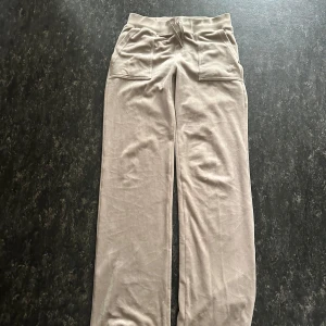 Beige mjukisbyxor Juicy Couture S - Beiga mjukisbyxor från Juicy Couture i mjuk velour med bred resår och snörning i midjan. Byxorna har stora fickor fram och bak, samt broderad logga på ena fickan. Perfekta för en chill och avslappnad stil.