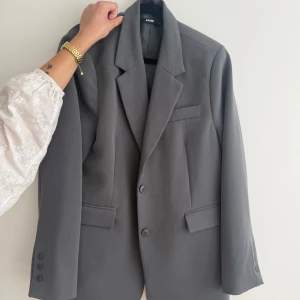 Grå kavaj från Bik Bok - Stilren grå kavaj från Bik Bok med klassisk krage, två knappar framtill och tre dekorativa knappar vid ärmslutet. Kavajen har två fickor med lock och en bröstficka. 