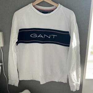 Vit sweatshirt från GANT med blå detalj - Vit sweatshirt från GANT med blått band och broderad logga över bröstet. Tröjan har rund hals, långa ärmar och ribbade muddar. Snygg kontrast med rosa linje på bröstet. Perfekt för en clean och sportig stil.