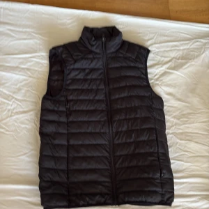 Svart dunväst från Livergy - Svart quiltad dunväst från Livergy med hög krage och hel dragkedja framtill. Västen har en stilren look med horisontella sömmar och två sidofickor. Perfekt att slänga över en hoodie för extra värme under kyliga dagar.