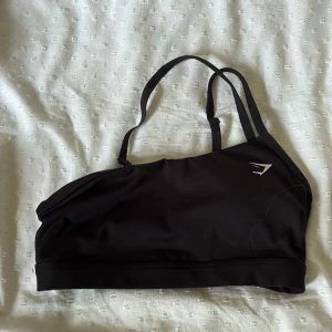 Svart one sholulder top från Gymshark - Säljer en svart top från Gymshark med smala justerbara axelband och diskret logga framtill. Toppen är i stretchigt material som passar perfekt till träning eller chill. Enkel och stilren design som är lätt att matcha med allt.