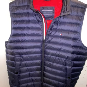 Mörkblå dunväst från Tommy Hilfiger - Snygg mörkblå dunväst från Tommy Hilfiger med röd insida och diskret logga på bröstet. Västen är quiltad med horisontella sömmar, har dragkedja framtill och två fickor med knappstängning. Perfekt för lager-på-lager-stil under kyliga dagar. Den är i Storlek L och passar M-L. Har du några funderingar tveka inte på att skriva😆