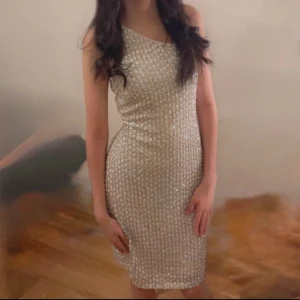 Glittrig one shoulder klänning - Snygg glittrig kortklänning i vit med ett tight passform och one shoulder-design. Klänningen har ett skimrande paljettmönster som fångar ljuset och ger en festlig look. Perfekt för dig som vill sticka ut på festen!