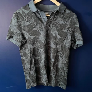 Grå pikétröja med blommigt mönster - Säljer en grå pikétröja från Dressmann i storlek S. Tröjan har ett snyggt blommigt och bladigt mönster i mörkare grå toner, klassisk krage och knäppning med två knappar. Perfekt för dig som vill ha en stilren men ändå unik look.