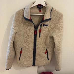 Beige fleecejacka från Patagonia - Säljer en beige fleecejacka från Patagonia med marinblå detaljer och röda dragkedjor. Jackan har hög krage, tre fickor med dragkedja och är perfekt för kyliga dagar. Mysig och stilren design som funkar till det mesta. Använt skick 