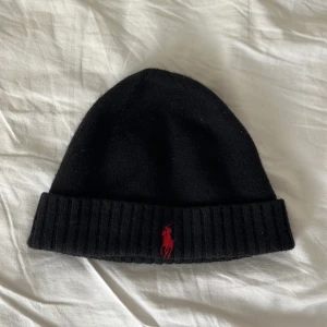 Svart mössa från Polo Ralph Lauren - Svart ribbstickad mössa från Polo Ralph Lauren med klassisk röd broderad logga framtill. Enkel och stilren design som passar till det mesta.