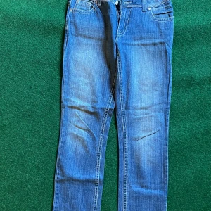 Blå jeans med broderade kors Jonna B - Snygga blå jeans från Jonna B i klassisk femficksmodell. Baksidan har broderade kors i vitt på bakfickorna som ger en unik look. Jeansen har normal passform och är tillverkade i slitstarkt denimtyg. Perfekta för dig som vill sticka ut lite extra.