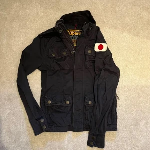 Svart jacka från Superdry med patch - Svart jacka från Superdry med fyra fickor framtill, knappar och dragkedja. Snygg japansk flaggpatch på ena ärmen och coola detaljer vid ärmslut. Perfekt för dig som gillar streetstyle och vill sticka ut lite extra.