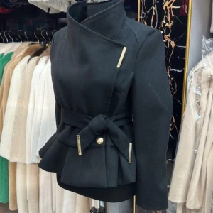 Svart kappa med bälte och detaljer - Stilren svart kappa med markerad midja och bälte med guldfärgade detaljer. Jackan har hög krage, asymmetrisk knäppning och peplum-liknande nederdel. Perfekt för dig som vill ha en elegant och trendig look.