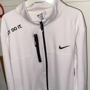 Vit Nike Dri-Fit vindjacka L - Snygg vit Nike Dri-Fit vindjacka med svarta detaljer och dragkedjor. Jackan har hög krage, tre fickor med dragkedja och texten 'JUST DO IT.' på axeln. Perfekt för dig som gillar sportig och clean stil.