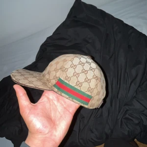 Gucci keps med GG-mönster och ränder - Snygg beige keps från Gucci med klassiskt GG-monogram över hela kepsen. Har gröna och röda ränder på sidorna som ger den en ikonisk look. Skärmen är böjd och materialet känns slitstarkt och lyxigt. Perfekt accessoar för att lyfta din outfit. Inga 1:1or, allt äkta. Pris kan diskuteras