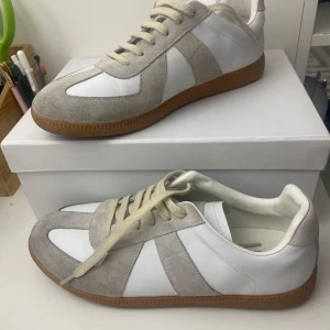 Maison Margiela sneakers vit - Maison Margiela sneakers i vitt skinn och ljusgrå mocka med klassisk gummisula. Snygg retrostil med rund tå och snörning. Perfekta för dig som gillar minimalistisk och stilren design.