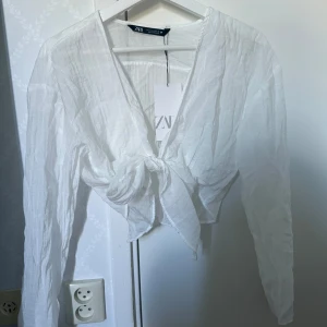 Vit omlottblus med knyt från Zara - Superfräsch vit omlottblus från Zara med lång ärm och snygg knytning framtill. Blusen är i ett lätt och luftigt tyg som ger en chill vibe och har en croppad passform. Perfekt för dig som gillar stilrena och trendiga plagg.