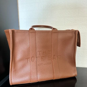 Brun Tote Bag från Marc Jacobs - Säljer en snygg brun Tote Bag från Marc Jacobs. Har logga framtill, två handtag och avtagbar axelrem med guldfärgade detaljer. Storleken är 42x32x16. Väskan kommer inte till användning längre, men har använt den bara några få gånger så väskan är i nyskick. Har tyvärr inga bevis på att väskan är äkta då jag har fått den i present.