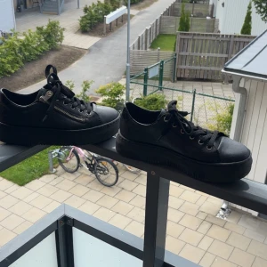 Svarta sneakers med dragkedja från Rieker - Snygga svarta sneakers från Rieker med chunky platt sula, snörning och cool dragkedjedetalj på sidan. Skorna är i skinn och har rund tå, perfekt för dig som gillar en stilren men edgy look.