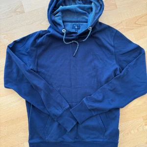 Marinblå hoodie från Gant - Snygg marinblå hoodie från Gant med klassisk huva och dragsko. Tröjan har en känguruficka framtill och är tillverkad i mjuk bomull. Perfekt för en chill och avslappnad stil. Långa ärmar och diskret Gant-logga på bröstet.