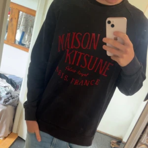 Maison Kitsuné svart sweatshirt - Svart sweatshirt från Maison Kitsuné med röd tryckt text på bröstet. Tröjan har klassisk rund halsringning, långa ärmar och är tillverkad i mjuk bomull. Perfekt för dig som gillar streetstyle och vill ha något stilrent men ändå med attityd.