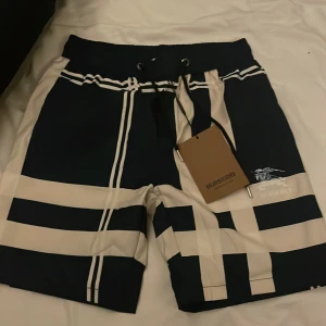 Burberry shorts - Snygga shorts från Burberry i marinblått och beige med breda ränder och coolt mönster. De har snörning i midjan och Burberry-logga framtill. Perfekta för sommaren och riktigt trendiga att bära till stranden eller stan.