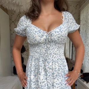 Blommig vit och blå kortklänning - Supersöt kortklänning i vitt med blått blommönster, smockad midja och puffärmar. Klänningen har v-ringning med snörning framtill och är tillverkad i ett mjukt bomullstyg. Perfekt för varma dagar och ger en romantisk vibe.