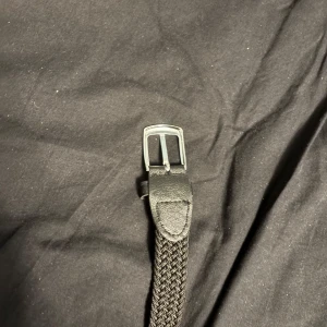 Svart flätat bälte med silverfärgat spänne - Snyggt svart bälte med flätad design och silverfärgat metallspänne. Bältet har detaljer i svart skinn vid spännet och är perfekt för att ge en stilren touch till din outfit. Passar både jeans och kostymbyxor.