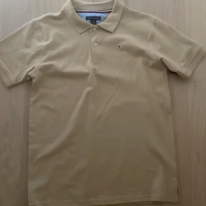 Beige pikétröja Tommy Hilfiger L - Klassisk beige pikétröja från Tommy Hilfiger i storlek L (14/16). Tröjan har korta ärmar, krage med två knappar och en liten broderad logga på bröstet. Tillverkad i mjuk bomull och perfekt för en clean och stilren look.