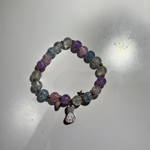 Pastellfärgat pärlarmband med kaninberlock - Sött armband med pärlor i pastellfärger som lila, rosa, blått och vitt. Armbandet har små guldfärgade detaljer och en gullig kaninberlock som hänger ner. Perfekt accessoar för dig som gillar lekfulla smycken med charmig touch.
