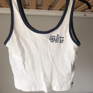 Vit ribbad linnetopp Pull&Bear - Vit ribbad linnetopp från Pull&Bear med marinblå kantband och broderad text på bröstet. Toppen har en croppad passform. Storlek S.