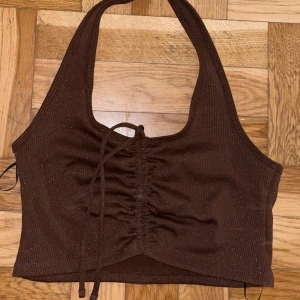 Brun ribbad halterneck topp S - Säljer en brun ribbad halterneck topp från Coco i storlek S. Toppen har snörning framtill som ger en snygg rynkad effekt och croppad passform. Perfekt för dig som gillar trendiga och bekväma plagg med lite edge.