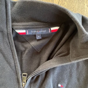 Svart half zip tröja Tommy Hilfiger L - Svart långärmad tröja från Tommy Hilfiger i storlek XL. Tröjan har en stilren half zip-dragkedja och diskret broderad logga på bröstet. Tillverkad i mjukt bomullsmaterial som känns skönt mot huden. Perfekt för en clean och klassisk look.